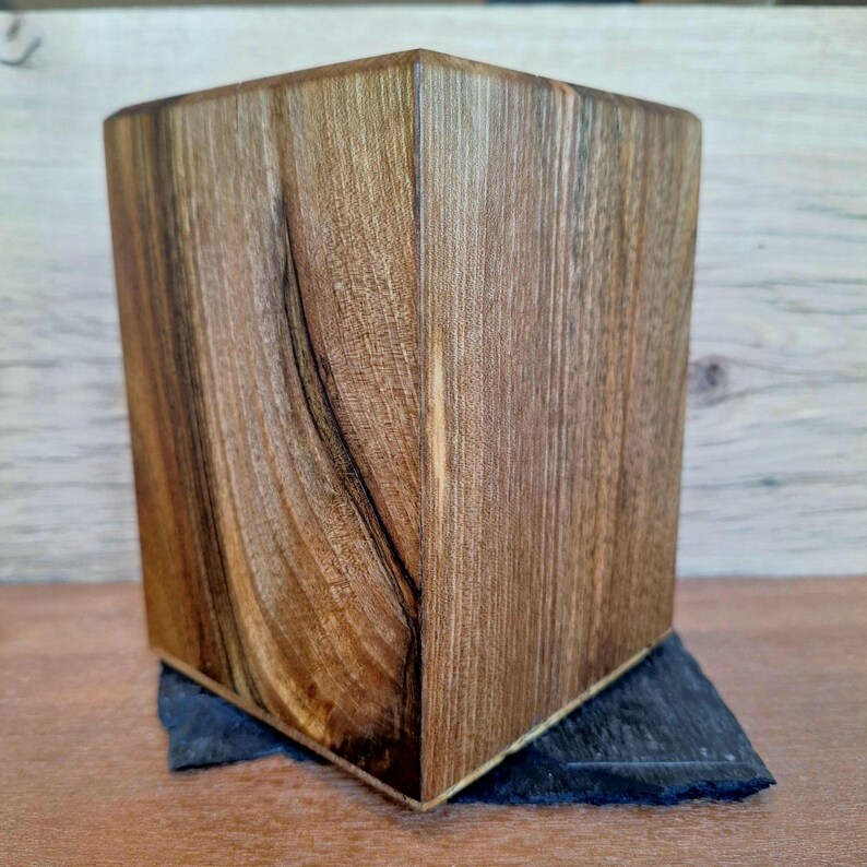 May include: A handcrafted wooden cube with a natural wood grain finish. The cube is set on a dark gray, triangular slate base. The wood has a warm brown tone, and the cube's edges are precisely cut.