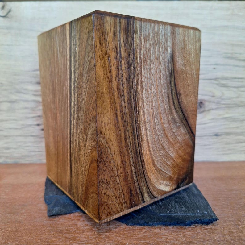 May include: A rectangular wooden box with a natural wood grain pattern. The box is a warm brown color and sits on a dark gray, irregularly shaped stone base. The wood grain has a swirling pattern.
