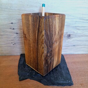 May include: A wooden pencil holder with a geometric design. The holder is made of dark brown wood and has a pencil inside. It sits on a black, irregularly shaped slate base.