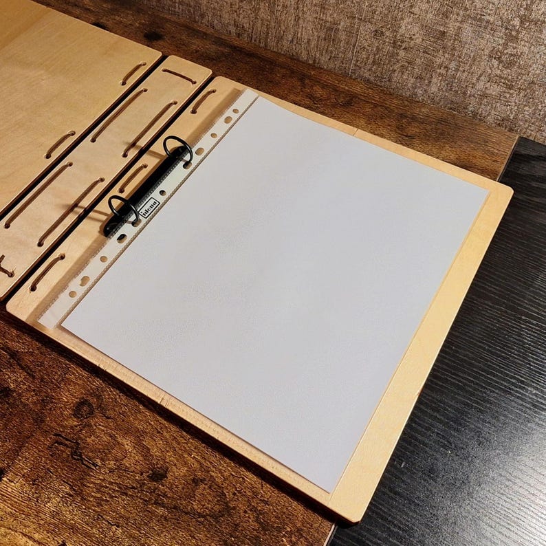 May include: A wooden binder with a light-colored wood frame and a black metal ring binder. The binder is open, revealing a white sheet of paper. The binder is on a wooden surface.