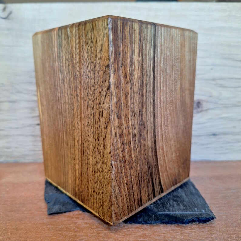 May include: A handcrafted wooden box with a geometric design. The box is made of dark brown wood with visible grain patterns. It sits on a dark gray, rectangular base, showcasing a natural, rustic aesthetic. The box has a square shape.