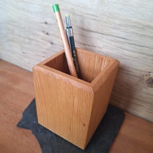 May include: A wooden pencil holder with a square shape and natural wood grain. Two pencils are inside the holder, one with a green tip and the other with a black body. The holder sits on a dark gray surface.