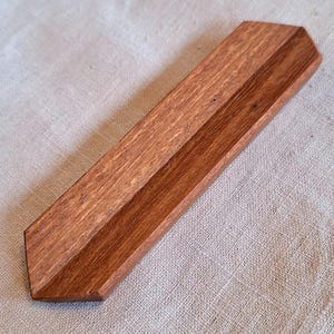 May include: A handcrafted wooden incense holder with a rich, reddish-brown finish. The rectangular tray features a central groove for holding incense sticks. The holder's angled edges and natural wood grain add to its rustic charm.
