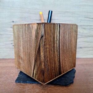 May include: A handcrafted wooden desk organizer with a geometric design. The organizer is made of dark brown wood with visible grain patterns and sits on a small, dark gray square base. Two pens are visible inside the organizer.