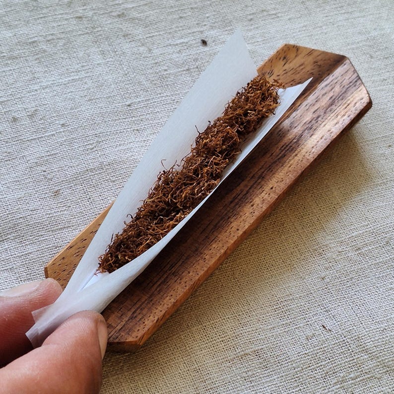 May include: A wooden rolling tray with a white paper and brown tobacco. The tray is rectangular with a groove to hold the paper and tobacco. The tobacco is loose and filling the paper.