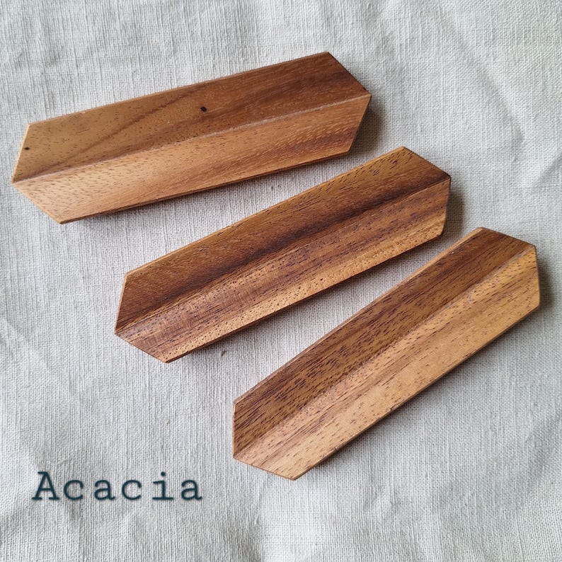 May include: Three acacia wood coasters with angled edges are arranged on a neutral fabric. The coasters are a warm brown color, showcasing the natural wood grain. The word "Acacia" is printed in black.
