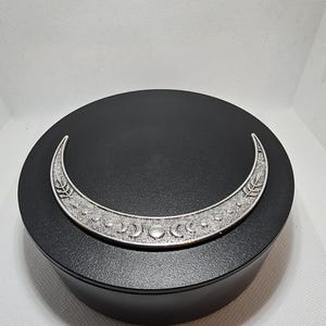 Pode incluir: Um porta-incenso prateado em forma de crescente lunar com um design detalhado. O suporte é colocado numa plataforma redonda preta. O design inclui padrões de folhas e pontos.