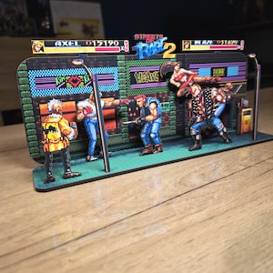 Puede incluir: Un diorama 3D multicapa del videojuego Streets of Rage 2 con personajes de pixel art de Axel y Blaze en un entorno de calle. La pantalla incluye una interfaz de juego superior con barras de salud y el logotipo del título en azul y rojo.