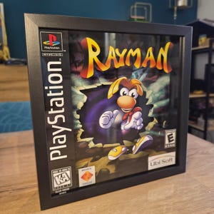 Puede incluir: Arte de la caja del juego Rayman de PlayStation enmarcado. La obra de arte presenta el título del juego en amarillo y naranja, con el personaje Rayman en el centro. El marco es negro y el logotipo de PlayStation es visible en el lateral.