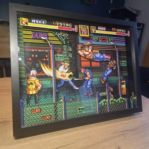 Peut inclure: Œuvre d'art 3D encadrée représentant une scène de combat de rue pixélisée d'un jeu vidéo classique. L'œuvre présente des personnages en action, avec des couleurs vives et une esthétique rétro. Le cadre est noir et l'œuvre est exposée sur une surface en bois.
