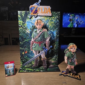 Peut inclure: Un ensemble de présentation Legend of Zelda Ocarina of Time comprenant une grande toile de fond verticale avec une illustration de personnage, une petite figurine assortie sur socle et un boîtier de jeu miniature. Le personnage porte une tunique verte.