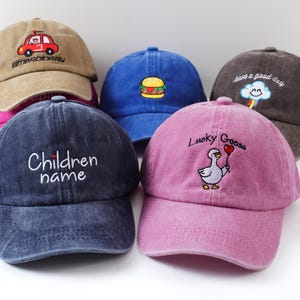 May include: A collection of six colorful baseball caps. The caps are in various colors, including brown, blue, and pink. Each cap features embroidered designs, such as a car, a burger, a cloud, a goose, and text that reads "Children name".