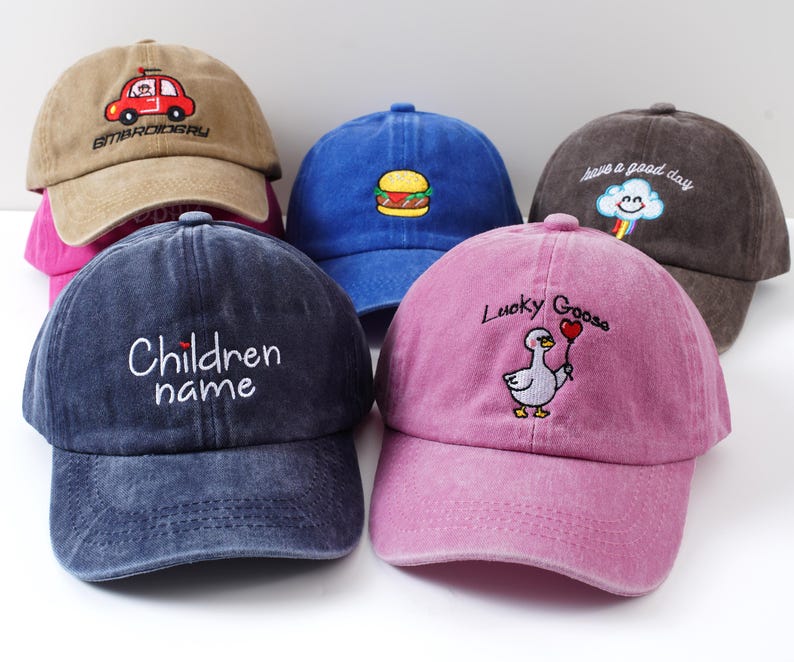 May include: A collection of six colorful baseball caps. The caps are in various colors, including brown, blue, and pink. Each cap features embroidered designs, such as a car, a burger, a cloud, a goose, and text that reads "Children name".