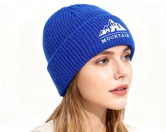 May include: A blue knit beanie with a white mountain graphic and the word "MOUNTAIN" in white. The hat is a solid blue color and has a folded brim. The beanie is made of a soft, ribbed knit material.