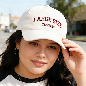 以下が含まれることがあります： クリーム色の野球帽。 深い赤色の文字で「LARGE SIZE CUSTOM」と書かれています。 つばはカーブしており、クラシックな6パネルのデザインです。 帽子は人が持っています。