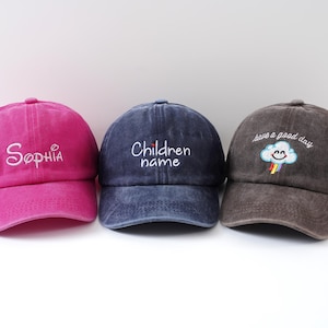 May include: Three baseball caps in a row. The first is magenta with "Sophia" embroidered in white. The second is navy blue with "Children name" embroidered in white. The third is brown with a smiling cloud and rainbow, and the text "have a good day".