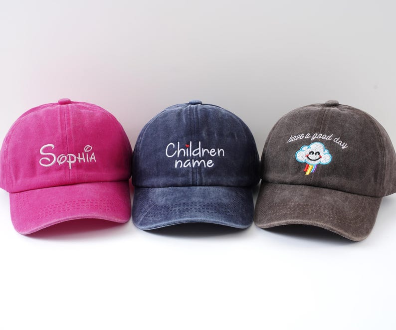 May include: Three baseball caps in a row. The first is magenta with "Sophia" embroidered in white. The second is navy blue with "Children name" embroidered in white. The third is brown with a smiling cloud and rainbow, and the text "have a good day".