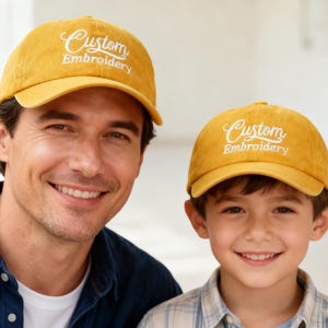 Op de afbeelding: Twee mensen dragen mosterdgele baseballpetten met "Custom Embroidery" in witte letters. De petten zitten op een man en een kind, beiden lachen naar de camera. De man draagt een blauw spijkeroverhemd en het kind draagt een geruit overhemd.