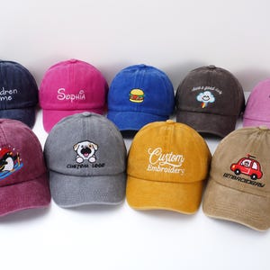 May include: A collection of colorful baseball caps with embroidered designs. The caps feature various designs, including a hamburger, a penguin, a dog, and text such as "Sophia" and "Custom Embroidery".