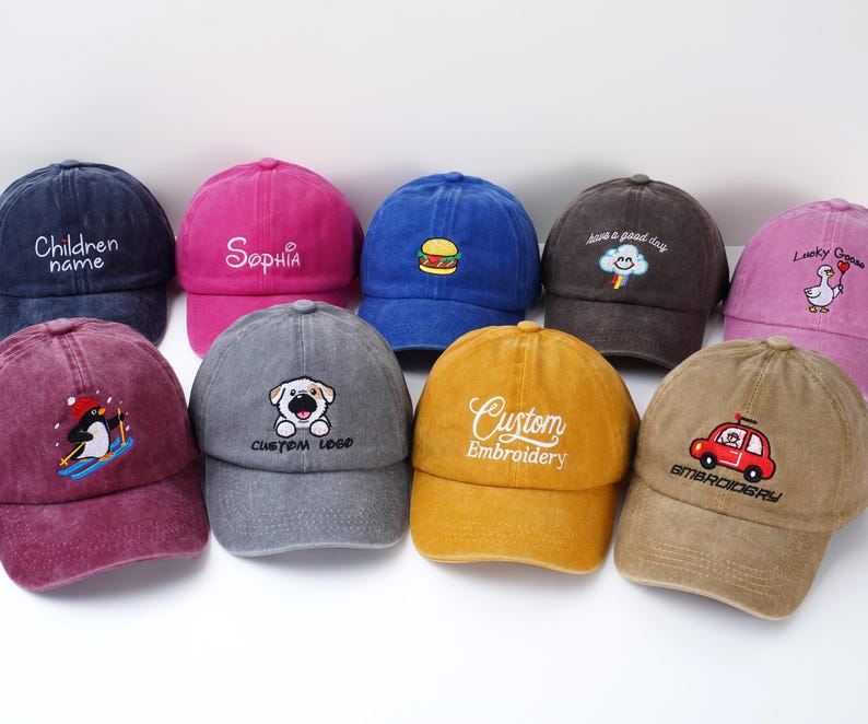 May include: A collection of colorful baseball caps with embroidered designs. The caps feature various designs, including a hamburger, a penguin, a dog, and text such as "Sophia" and "Custom Embroidery".