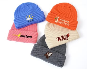 May include: A stack of six colorful knit beanies. The beanies are blue, pink, orange, cream, and gray. Each beanie has an embroidered design or text, including a moon and stars, a rabbit, and the word "WILD".