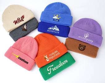 May include: A collection of colorful knit beanies in various colors, including cream, blue, pink, orange, purple, and green. Each beanie features embroidered designs and text, such as a wolf, mountain, lightning bolt, bear, and the words "WILD", "MOUNTAIN", "custom", "Freedom", and "accompany".