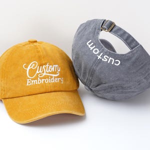 May include: Two baseball caps, one mustard yellow and one gray. The yellow cap has "Custom Embroidery" embroidered in white script. The gray cap has "custom" embroidered in white on the back. Both caps have adjustable straps.