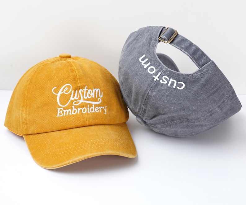 May include: Two baseball caps, one mustard yellow and one gray. The yellow cap has "Custom Embroidery" embroidered in white script. The gray cap has "custom" embroidered in white on the back. Both caps have adjustable straps.