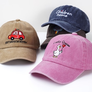 May include: Four baseball caps in various colors. One tan cap features a red car embroidery with the word "EMBROIDERY". A navy blue cap has the text "Children name". A pink cap displays a goose holding a heart balloon with the words "Lucky Goose".