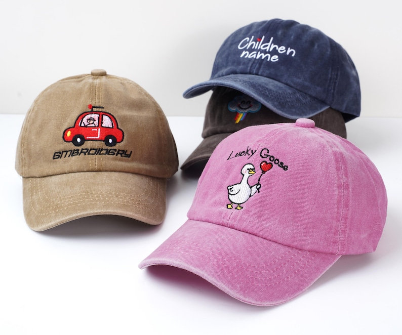May include: Four baseball caps in various colors. One tan cap features a red car embroidery with the word "EMBROIDERY". A navy blue cap has the text "Children name". A pink cap displays a goose holding a heart balloon with the words "Lucky Goose".