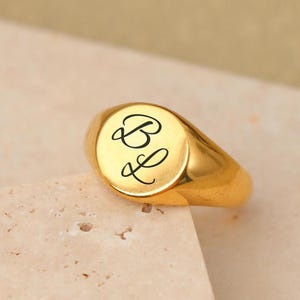 Peut inclure: Une bague chevalière dorée présentant une surface lisse et polie avec les lettres B et L gravées en écriture cursive noire sur la face circulaire. La bague possède un anneau solide et un design à sommet plat et arrondi avec une finition métallique.