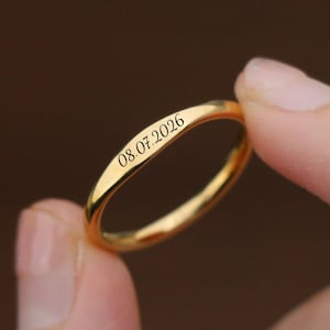 Könnte beinhalten: Ein goldfarbener Ring mit dem eingravierten Datum "08.07.2026". Der Ring wird zwischen zwei Fingern vor einem verschwommenen Hintergrund gehalten. Der Ring hat ein schlichtes Banddesign.