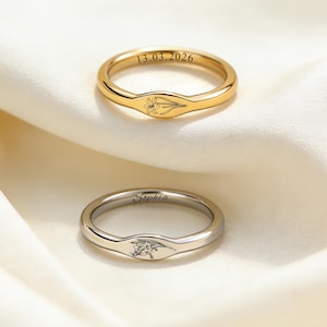 Puede incluir: Dos anillos, uno dorado y otro plateado, descansan sobre una tela de satén blanco. El anillo dorado tiene grabada la fecha "13.03.2026" y un diseño floral. El anillo plateado está grabado con el nombre "Sophia" y un pequeño grupo de piedras.