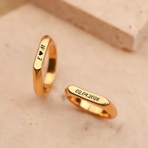 Può includere: Due anelli color oro con fasce rettangolari. Un anello ha incise le iniziali "E ♡ M". L'altro anello ha incisa la data "02.04.2026". Gli anelli sono esposti su una superficie di colore chiaro.