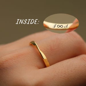 Op de afbeelding: Een goudkleurige ring met een gedraaid bandontwerp. Een ovale grafische inzet toont de binnenkant van de ring met de gegraveerde tekst INSIDE: gevolgd door de letters J en A gescheiden door een oneindigheidssymbool in een cursief lettertype.