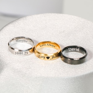 Puede incluir: Tres anillos con texto grabado. Un anillo plateado con "Marryme" y "10.01.25". Un anillo dorado con "Love Always" y "Lucy ❤ Tom". Un anillo negro con "David".