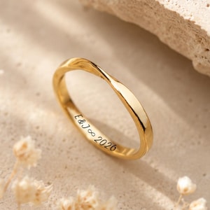 Peut inclure: Une bague dorée avec un design de bande torsadée. La surface intérieure de la bague est gravée avec le texte E&J∞2026 dans une police script noire. La bague présente une finition lisse et polie ainsi qu'une forme fluide et continue.