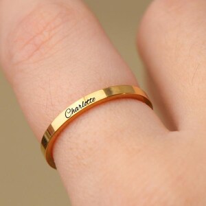 Könnte beinhalten: Ein goldfarbener Ring mit dem eingravierten Namen "Charlotte" auf dem Band. Der Ring hat ein schlichtes, flaches Banddesign und wird am Finger getragen. Der Ring besteht aus einem glänzenden, reflektierenden Metall.