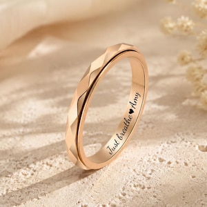 May include: A rose gold-toned ring with a faceted, geometric outer band surface. The inner band features the engraved black text Just breathe with a small heart symbol followed by the name Amy, clearly visible in the centre of the ring.