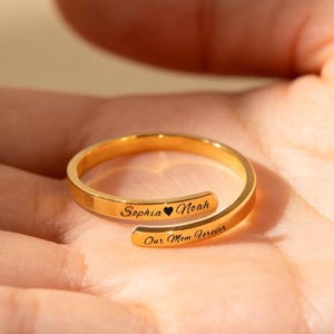 Könnte beinhalten: Ein goldfarbener Wickelring mit eingraviertem Text. Der Ring ist offen, wobei sich die Enden überlappen. Der Text auf dem Ring lautet "Sophia ❤ Noah" und "Our Mom Forever". Der Ring wird auf einer Hand gezeigt.