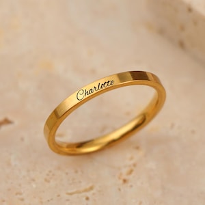 May include: A gold-colored ring with the name "Charlotte" engraved on the band. The ring is set against a neutral-toned background. The ring appears to be a thin band, likely for everyday wear.