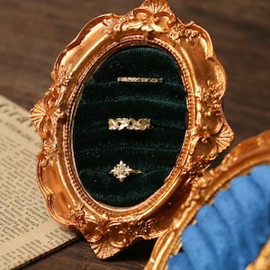 Könnte beinhalten: Ein goldfarbener, verzierter Rahmen mit ovaler Öffnung, der drei Ringe auf einer dunkelgrünen Samtoberfläche zeigt. Die Ringe weisen filigrane Designs auf, darunter florale und geometrische Muster. Die filigranen Details des Rahmens und die Eleganz der Ringe schaffen eine luxuriöse Ästhetik.