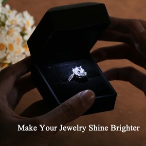 K&ouml;nnte beinhalten: Ein Diamant-Verlobungsring in einer schwarzen Samtbox, beleuchtet von einem kleinen Licht. Der Ring hat einen gro&szlig;en Mittelstein, umgeben von kleineren Diamanten. Der Text auf dem Bild lautet: "Make Your Jewelry Shine Brighter."