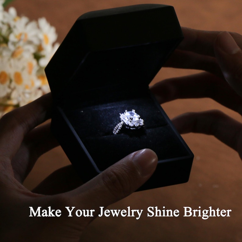 K&ouml;nnte beinhalten: Ein Diamant-Verlobungsring in einer schwarzen Samtbox, beleuchtet von einem kleinen Licht. Der Ring hat einen gro&szlig;en Mittelstein, umgeben von kleineren Diamanten. Der Text auf dem Bild lautet: "Make Your Jewelry Shine Brighter."