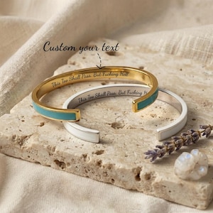 May include: Two open cuff bracelets, one gold-toned with a turquoise inlay and one silver-toned with a white inlay. Both feature the engraved text "This Too Shall Pass. But Fucking Hell" on the inner band. The text "Custom your text" is visible.