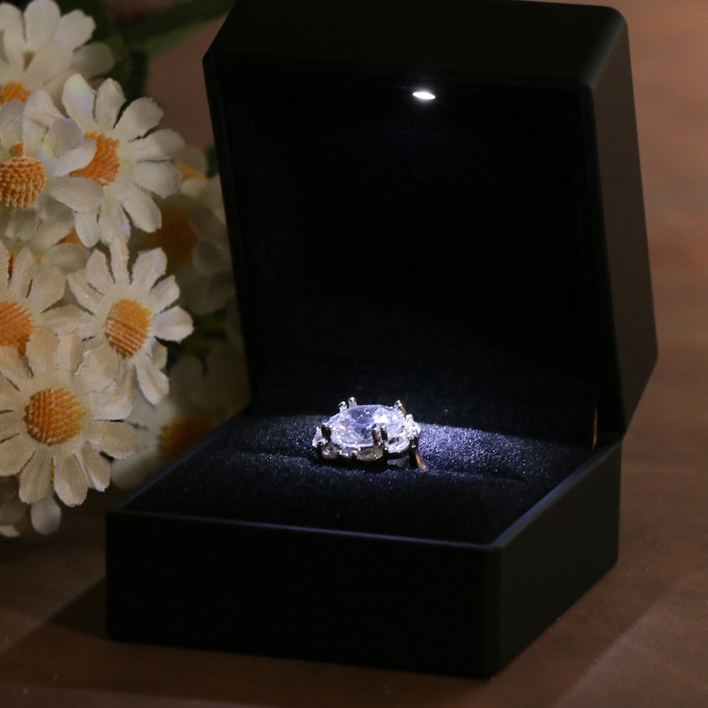 K&ouml;nnte beinhalten: Eine schwarze Ringbox mit einem Scheinwerfer, der einen silbernen Ring mit einem gro&szlig;en, klaren Edelstein beleuchtet. Der Ring liegt auf einem dunklen Samtinnenraum, mit wei&szlig;en Blumen im Hintergrund.