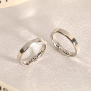以下が含まれることがあります： 平らで面取りされたデザインの2つのシルバーの結婚指輪。各リングの内側には日付「03.10.2025」が刻印されています。リングは白く、質感のある表面に表示されています。