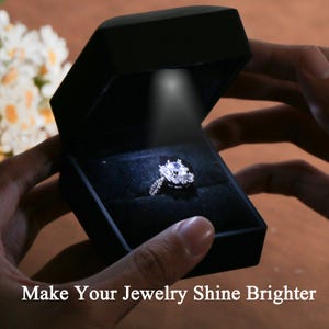 Könnte beinhalten: Eine schwarze Schmuckschatulle mit einem Scheinwerfer, der einen Diamantring beleuchtet. Der Ring hat einen großen, klaren Mittelstein, der von kleineren Steinen umgeben ist. Der Text "Make Your Jewelry Shine Brighter" steht unten.