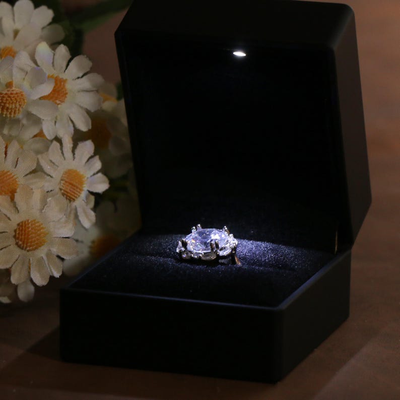 K&ouml;nnte beinhalten: Eine schwarze Ringbox mit einem Scheinwerfer, der einen silbernen Ring mit einem gro&szlig;en, klaren, oval geformten Stein beleuchtet. Der Ring ist mit kleineren Steinen um den Hauptstein besetzt. Die Box ist ge&ouml;ffnet.