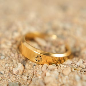 Può includere: Un anello dorato con una fascia piatta e un motivo a forma di sole nero. L'anello è posto su un letto di piccoli ciottoli beige, che ne evidenziano la lucentezza metallica e il design semplice. Un gioiello.